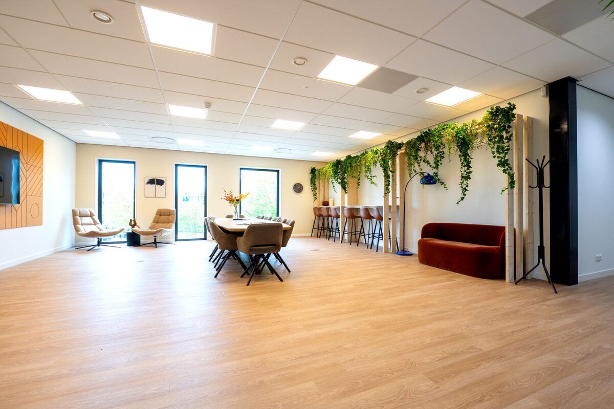 Lichte gemeenschappelijke ruimte in Enerzjyhûs met houten vloer, zithoek, bar en hangende groene beplanting.