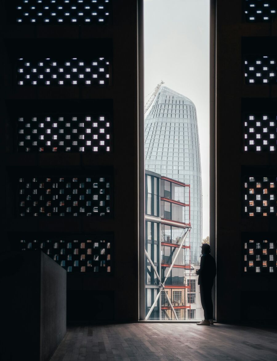 Een figuur staat bij een hoog, smal raam en kijkt uit over de stad — daglicht, ingetogen en koel van toon.